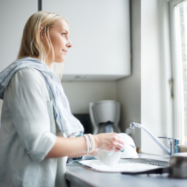 A blonde woman stands washing dishes looking out the window.