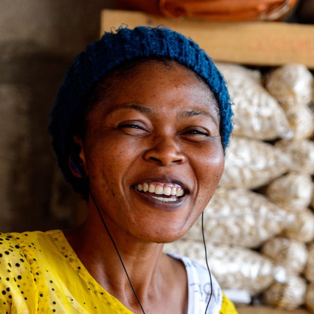 Ghanaian woman in yellow dress and blue hat wearing earbuds smiles in a stall with bagged produce behind her on shelves at the Kumasi market in Ghana.