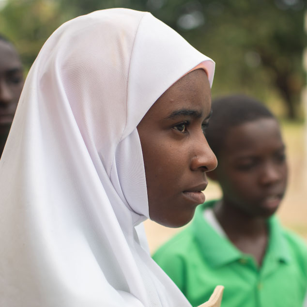 Adolescent female student wearing a white head covering in Dodma, Tanzania with a male student out of focus behind her wearing green polo shirt.